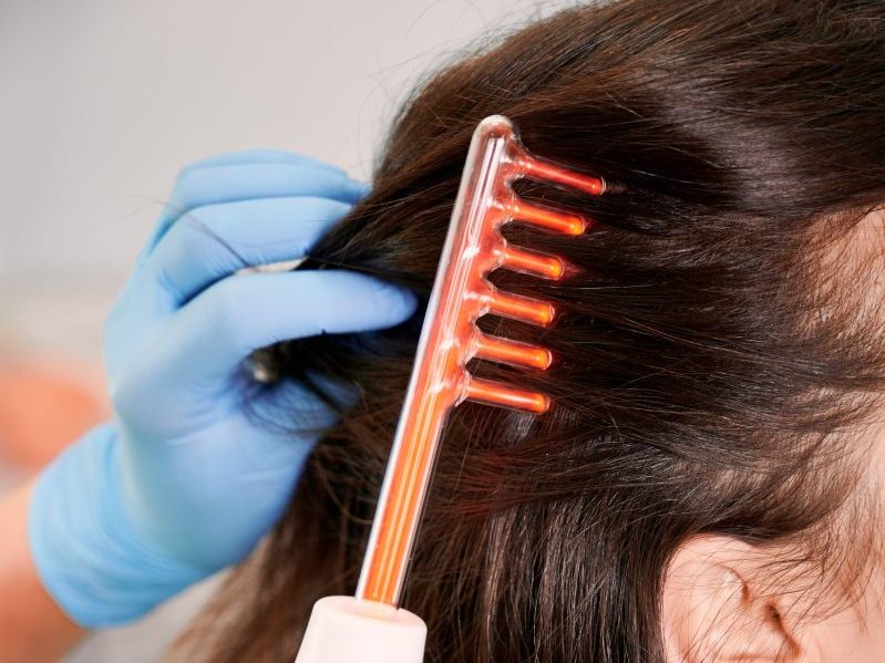 Close up of doctor trichologist hand in sterile gloves brushing female client hair with laser hair growth device. Woman receiving hair treatment laser therapy in medical center. Concept of hair care.